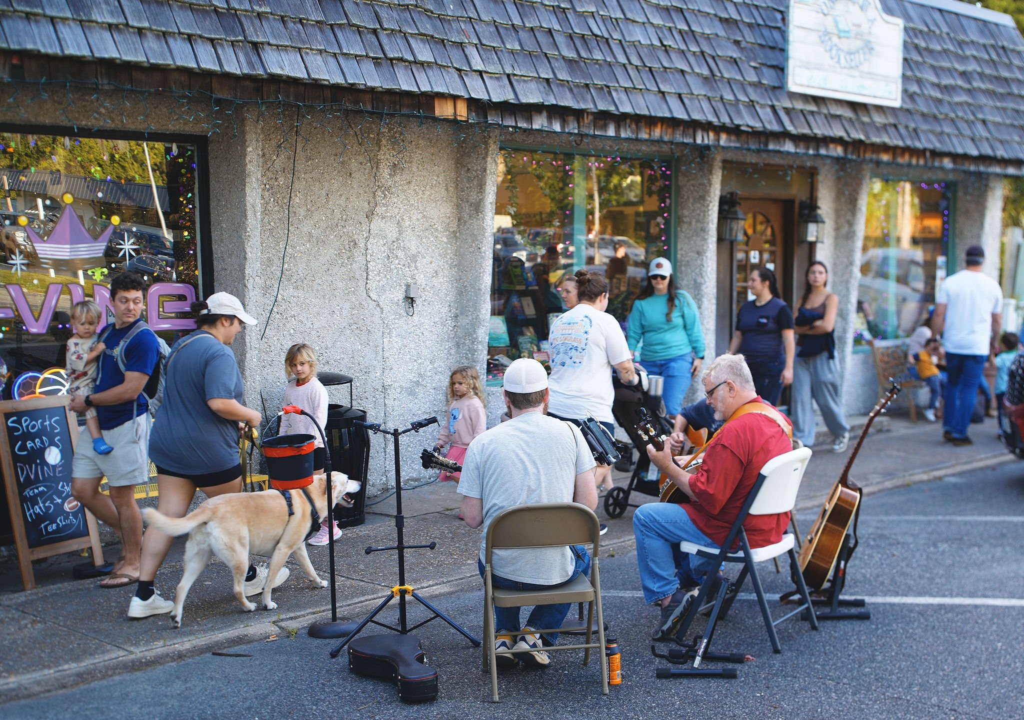 People gathering outside a storefront, some are playing guitar while others are watching. Children and a dog are present.