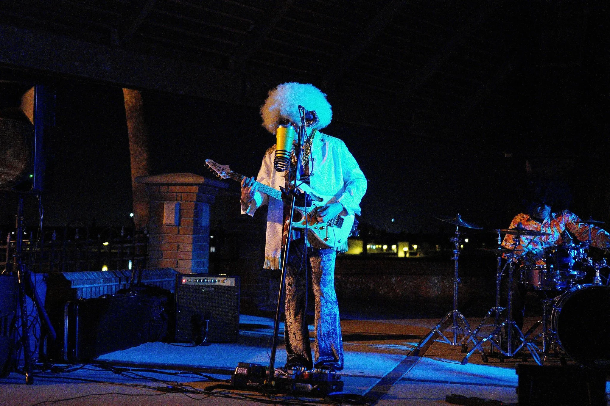 A musician with curly white hair and glasses performs on stage at night, playing an electric guitar and singing into a microphone, with a drummer in the background. The stage has musical equipment and is outdoors against a cityscape.