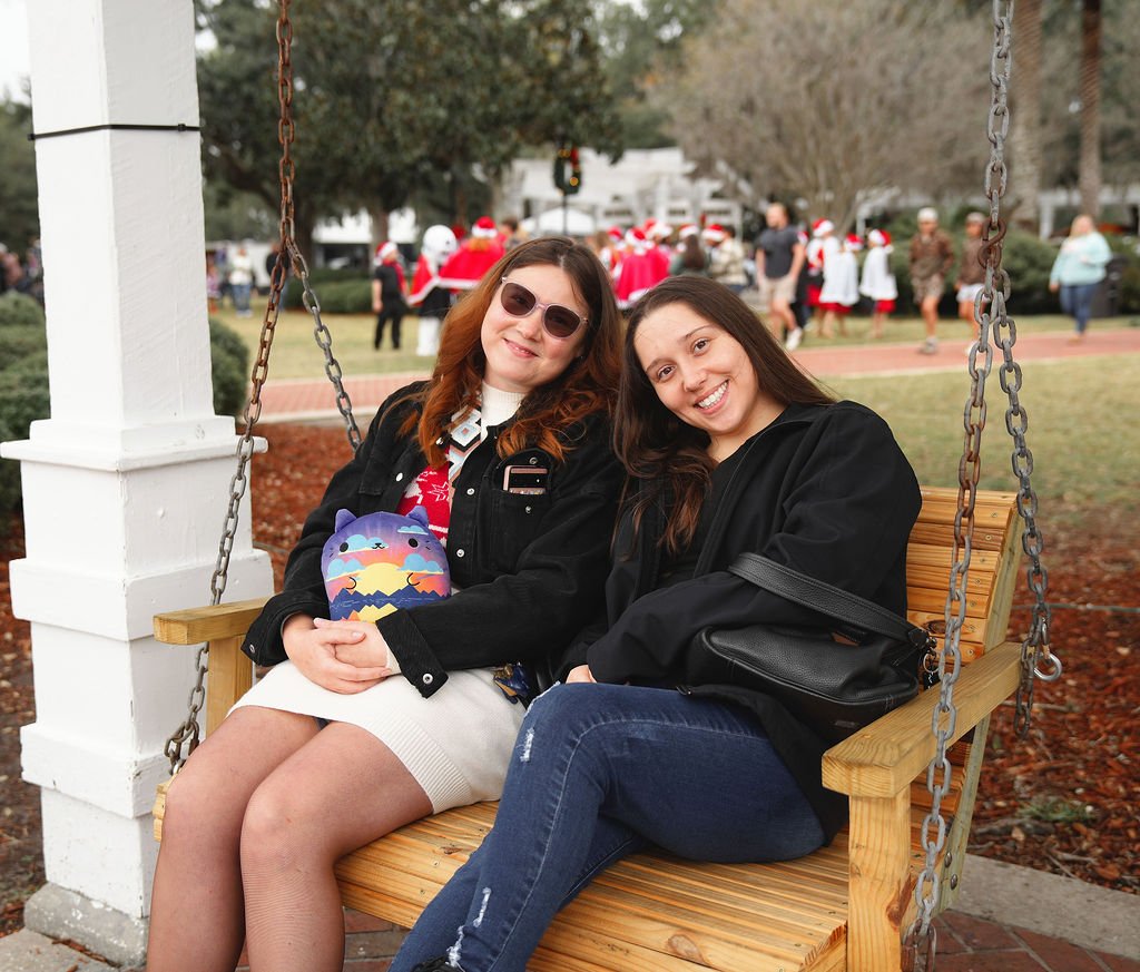 Two young women sitting on a wooden swing bench outdoors, smiling at the camera, with Christmas-themed event in the background, including people wearing Santa hats.