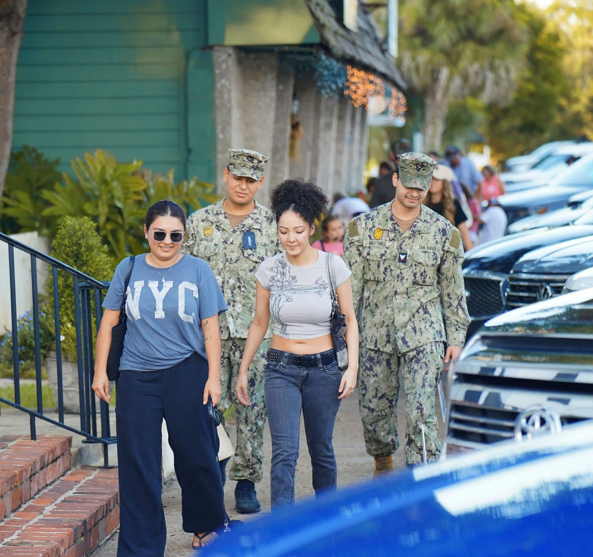 Group of young women and U.S. Navy soldiers walking outdoors on a sidewalk, with parked cars nearby, during daytime.