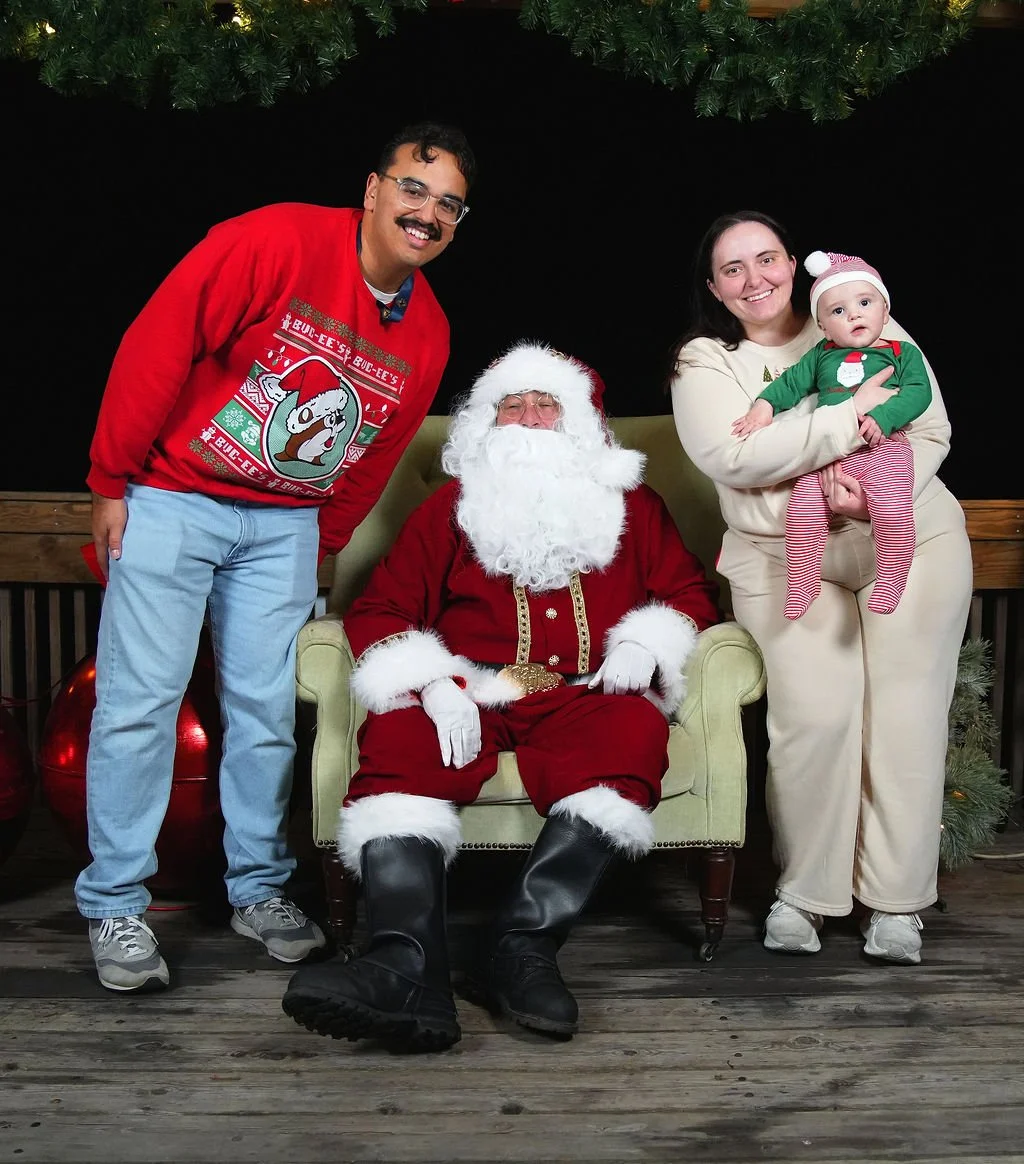 People dressed in Christmas attire posing with Santa Claus, sitting on a green chair, with Christmas decorations around.