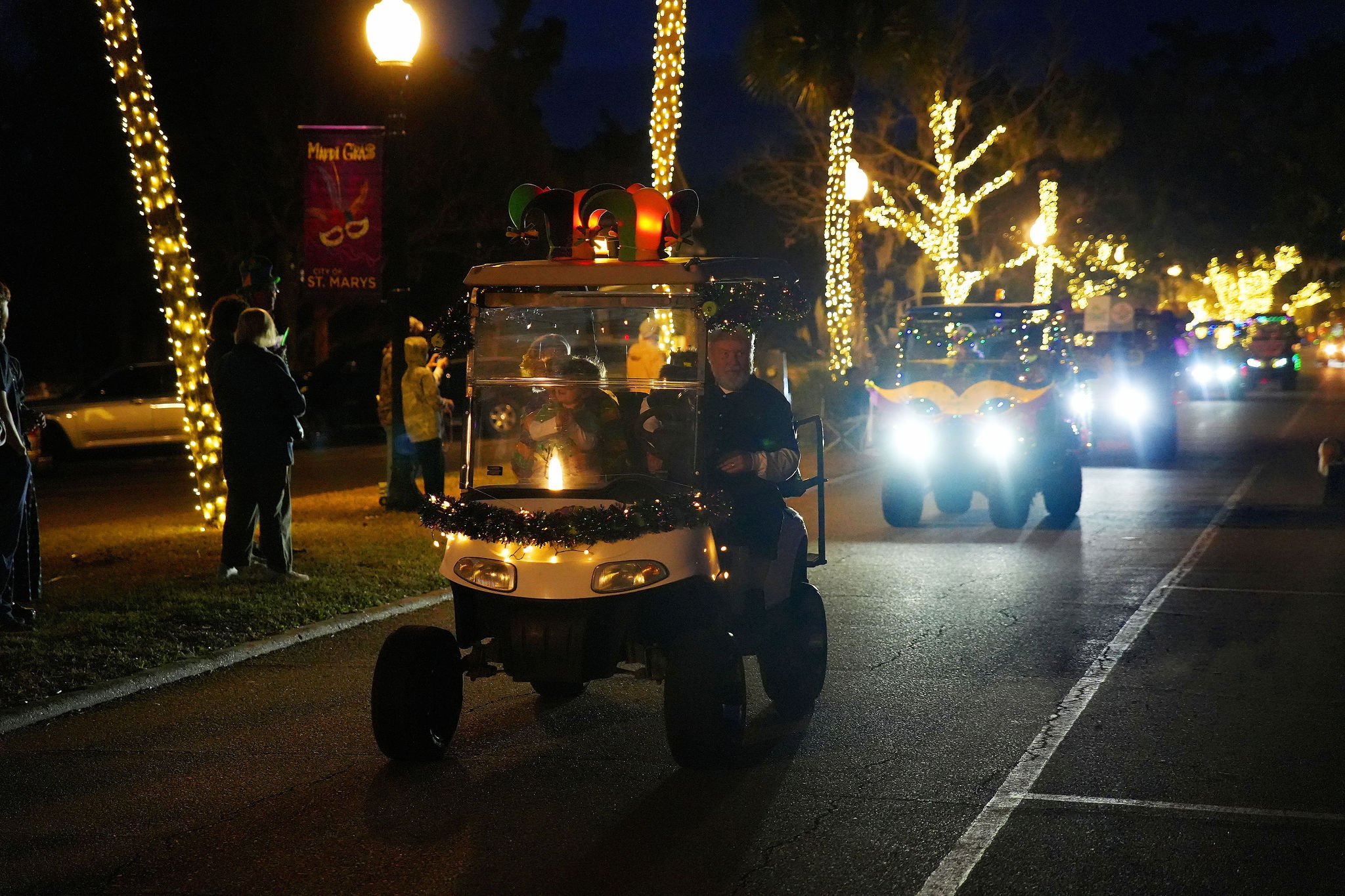 A nighttime parade scene with decorated golf carts and people watching. The golf carts are adorned with Christmas lights and ornaments, and some have Christmas-themed decorations. The street is lined with lit trees, and people are standing along the 
