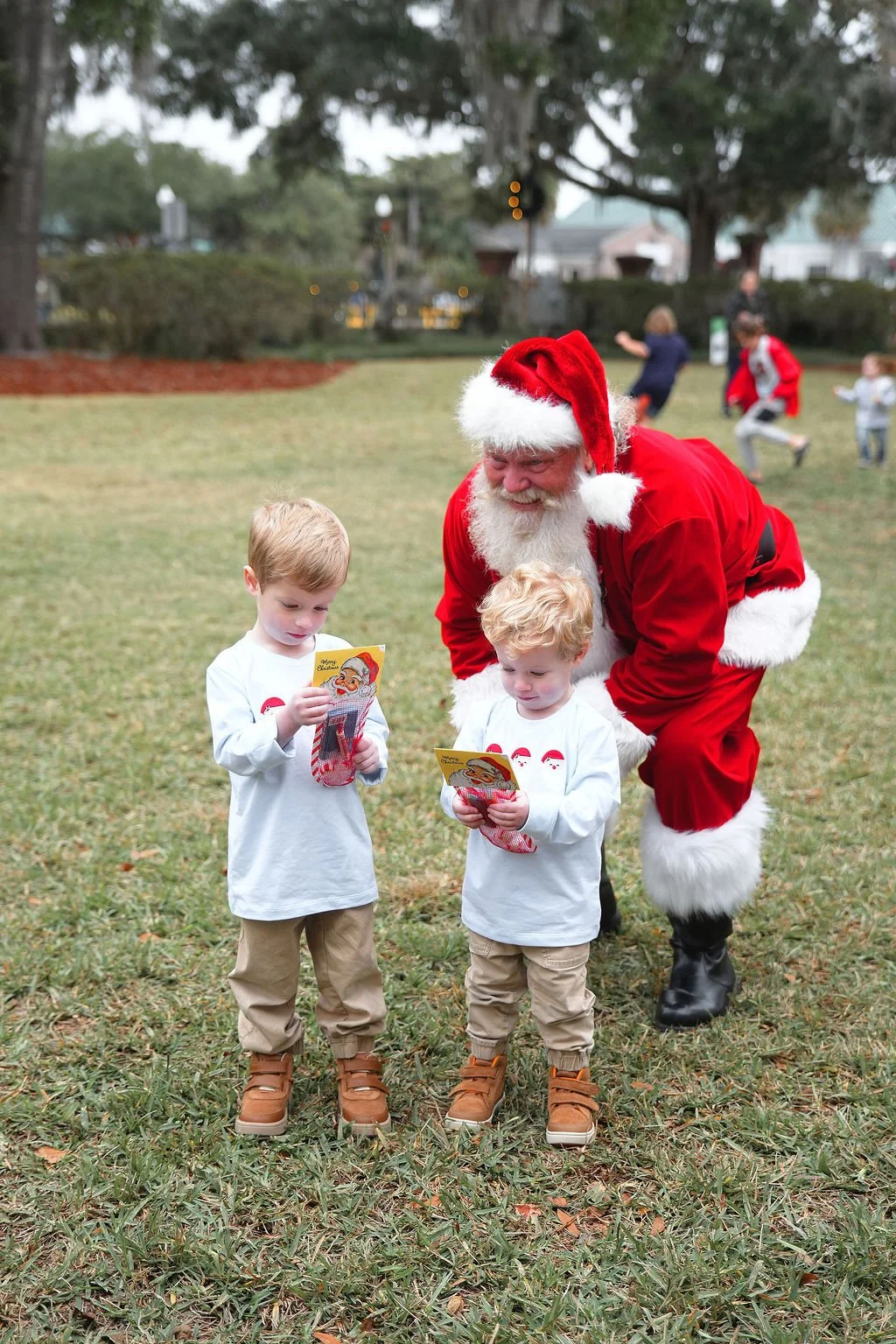 Two young boys with light skin and blond hair reading holiday cards while Santa Claus, dressed in a red suit with a white beard, squats behind them on a grassy outdoor area. In the background, other children and people are playing near large trees an