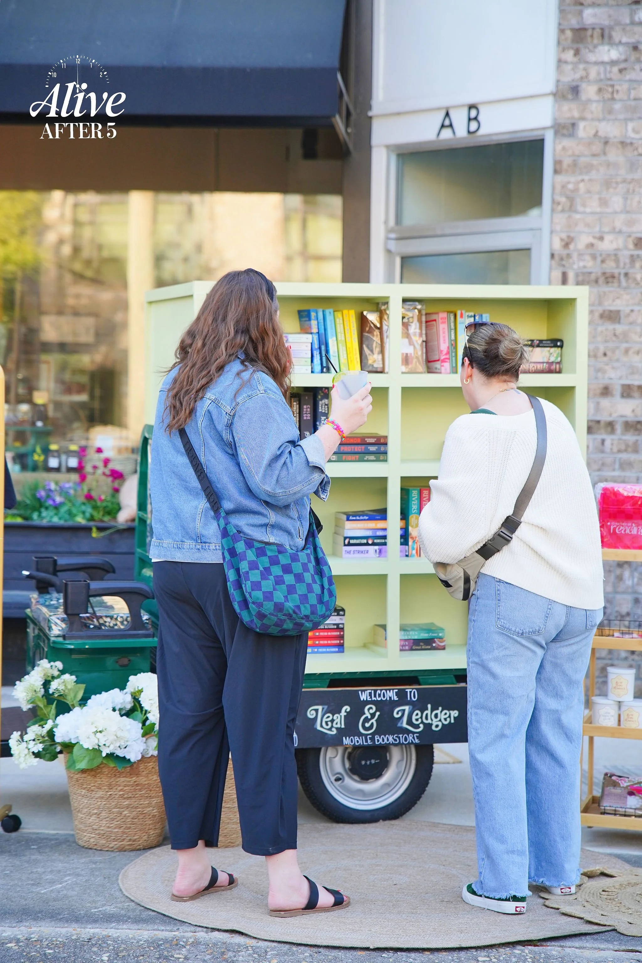 Two women browsing books at a mobile bookstore called Leaf & Ledger, set up on a sidewalk.