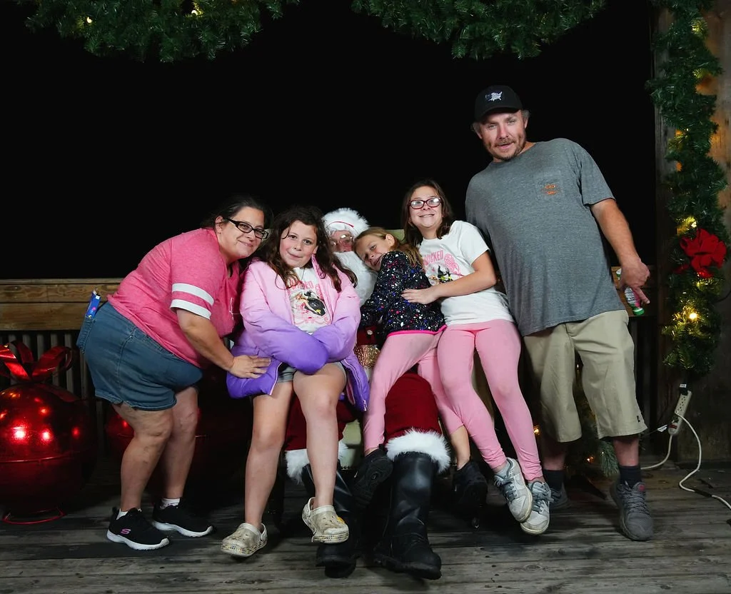 Group of six people, including children and adults, posing with Santa Claus on a wooden stage decorated for Christmas. The background has a Christmas tree and lights.