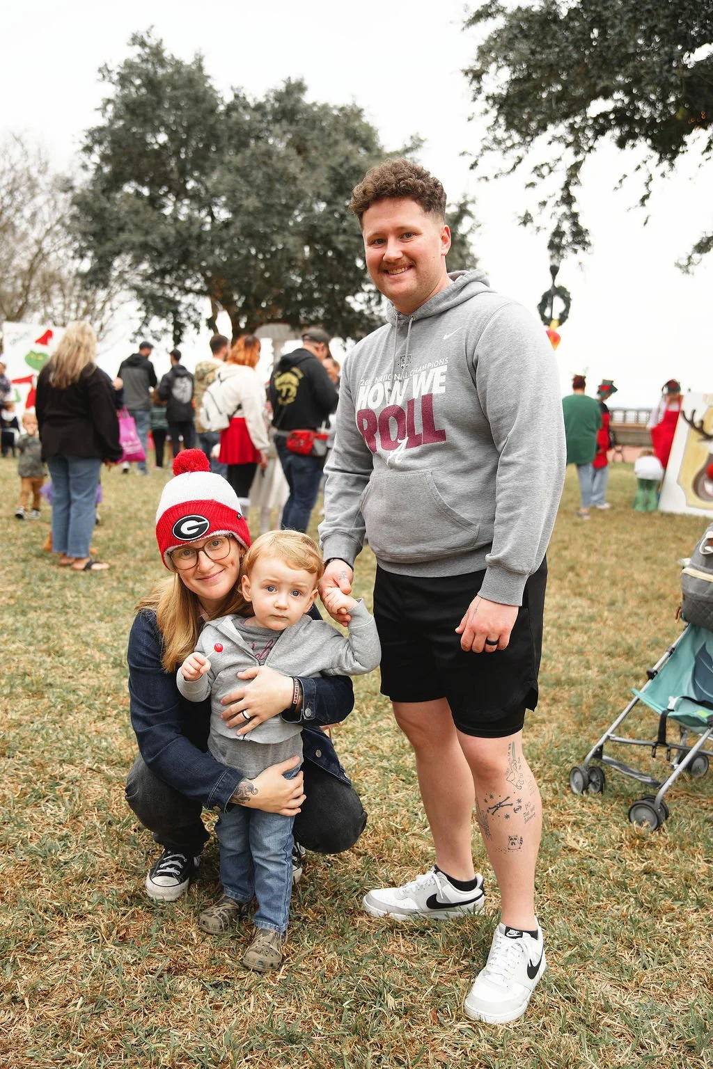 A smiling man, woman, and young child posing outdoors at a festive event with trees and other people in the background. The woman is wearing a Georgia Bulldogs beanie and glasses, and is kneeling, holding the young child. The man is standing, holding