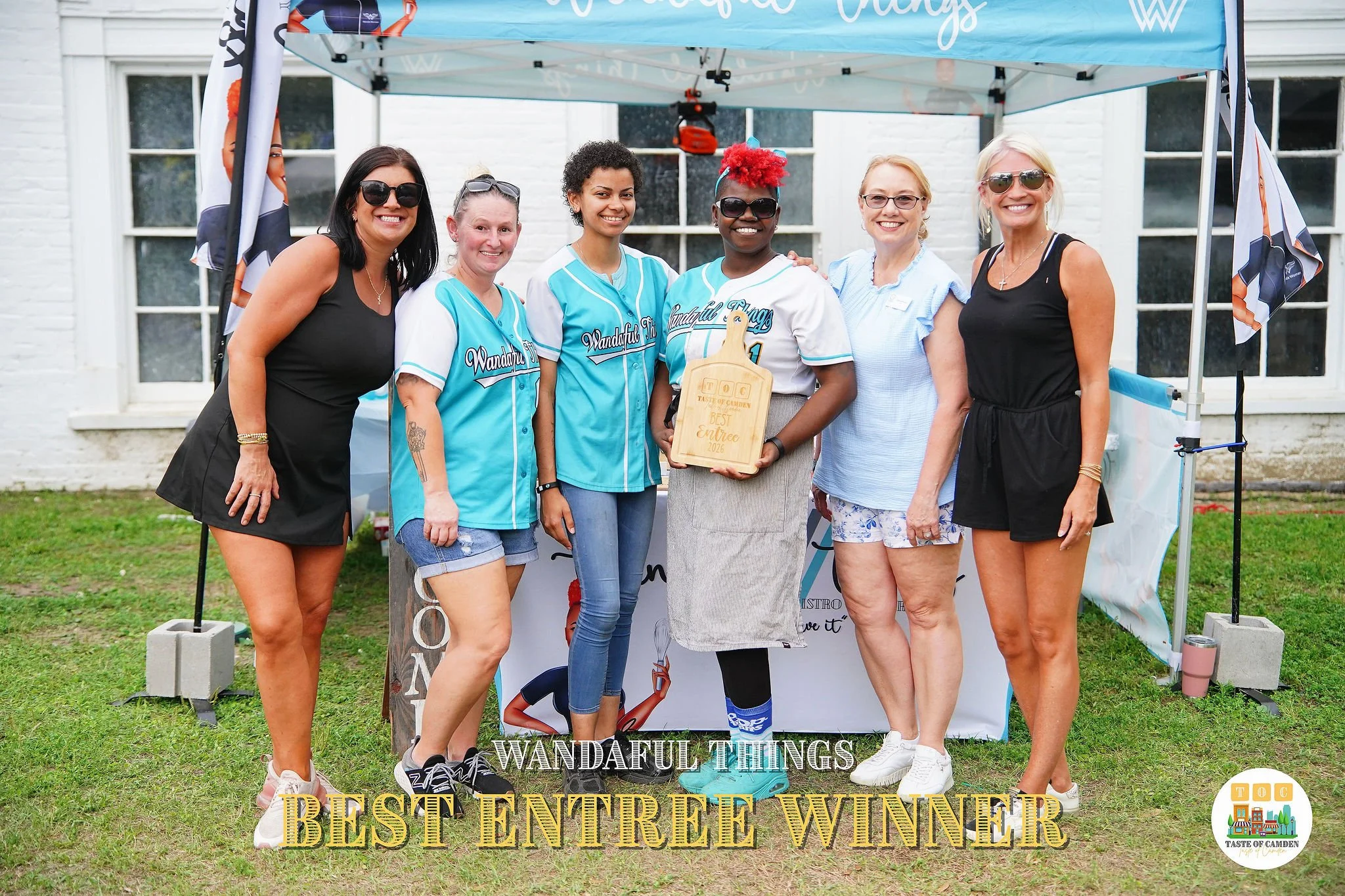 Group of six women standing outdoors under a Canopy, posing for a photo. The woman in the middle holds a wooden cutting board with the words 'TOC Taste of Camden Best Entrée 2022.' The women are smiling, and there are two flags on either side of the 