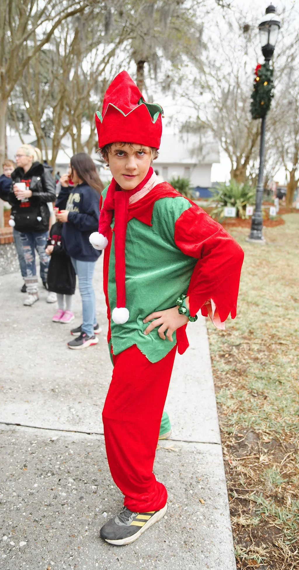 A young boy dressed as an elf with a red and green costume, red hat with a green trim, standing outdoors on a sidewalk with other people in the background.