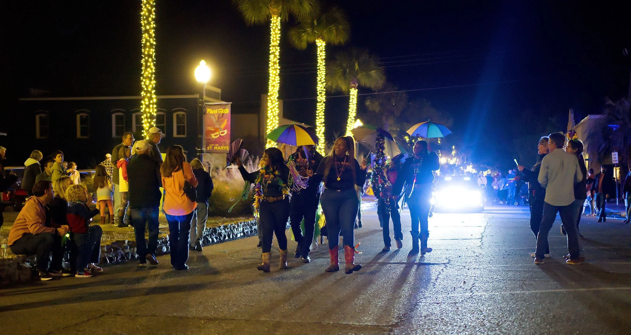 Nighttime street scene with people celebrating, some holding umbrellas, dressed in festive attire, and walking or standing in groups. Bright lights illuminate the area, with palm trees wrapped in string lights and a police car with flashing lights in
