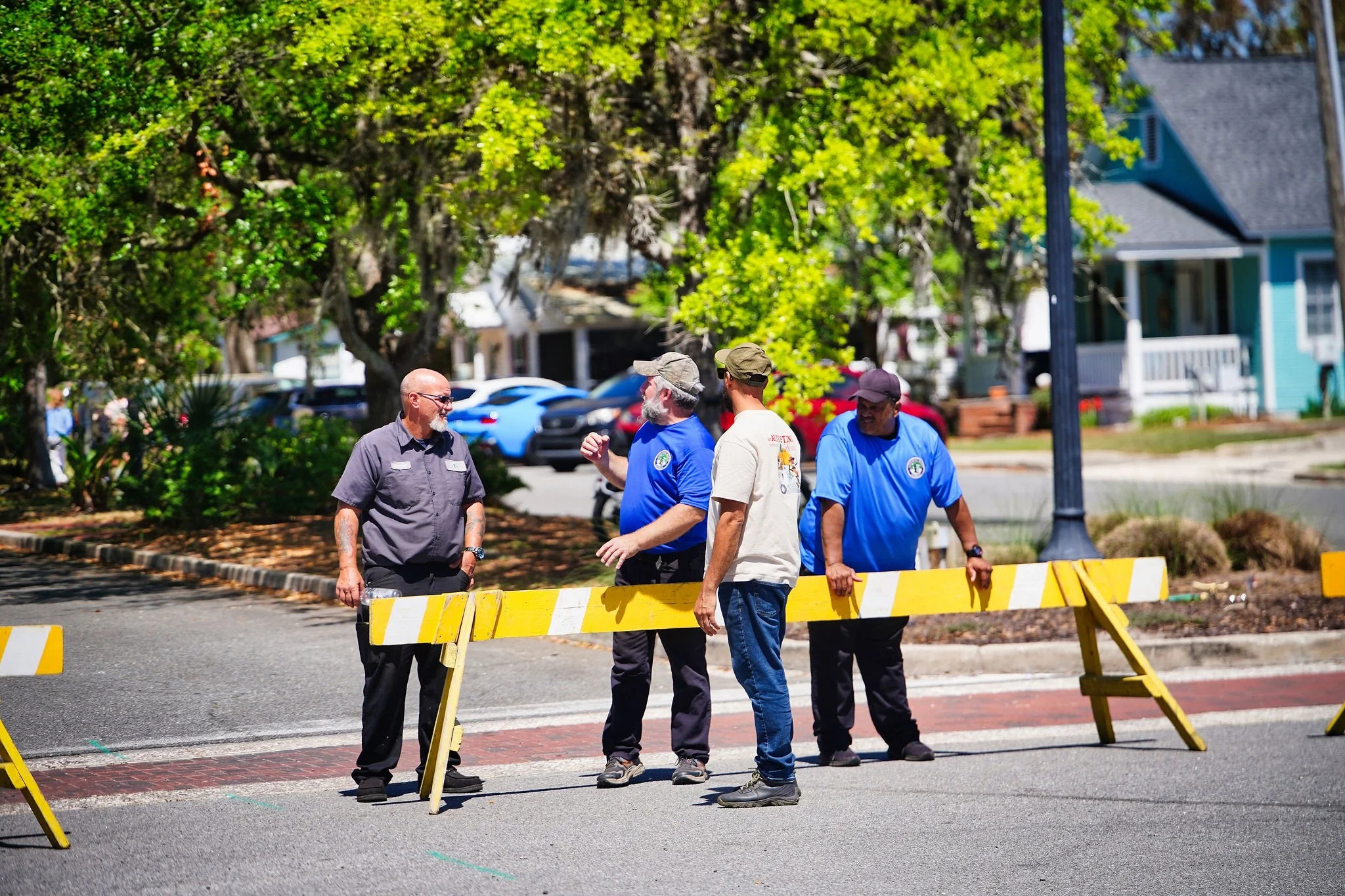 Group of five men standing behind yellow and white barricades on a street, having a conversation during daytime with trees and houses in the background.