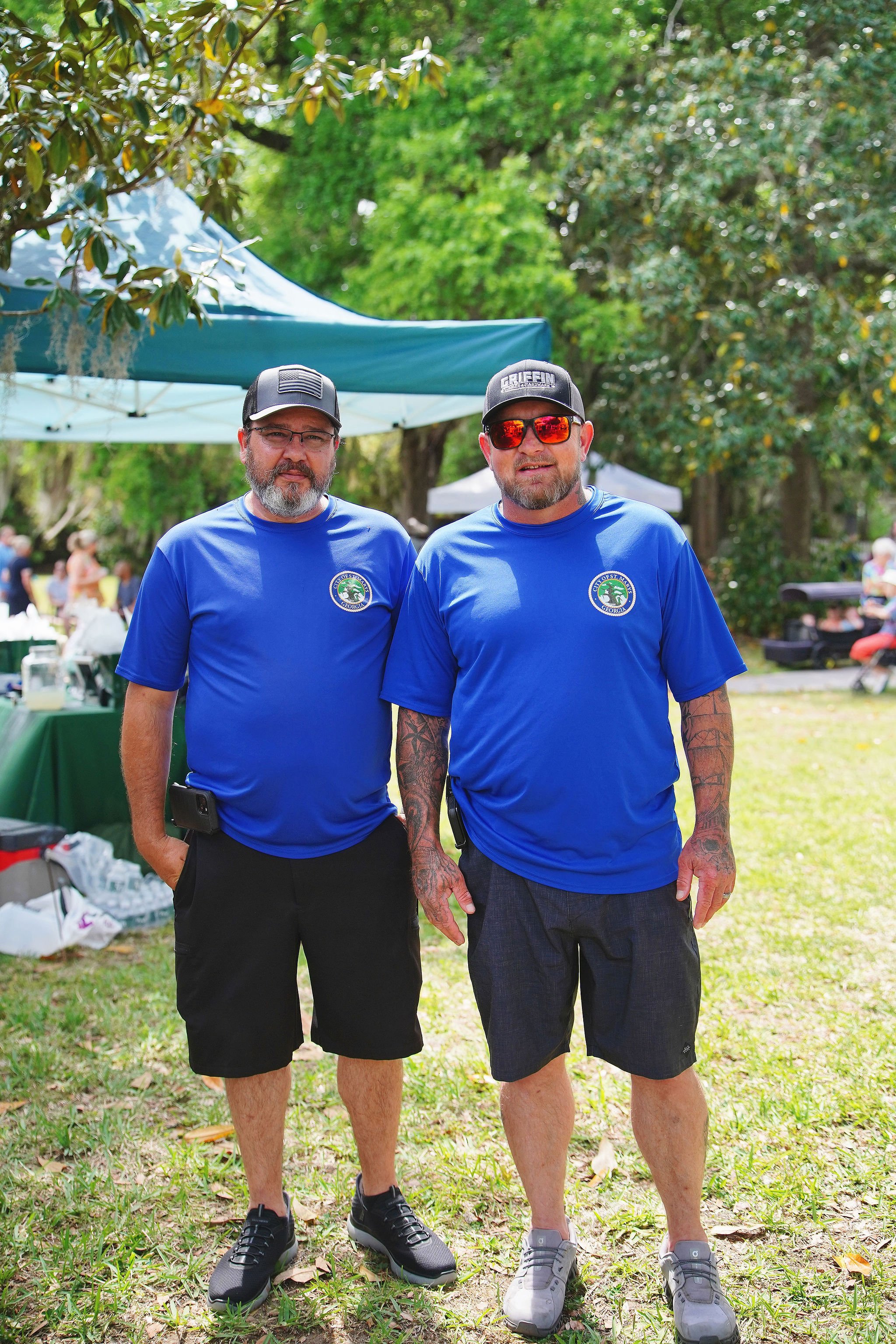 Two men standing outdoors on grass, wearing matching blue shirts with a logo, with trees and a tent in the background.