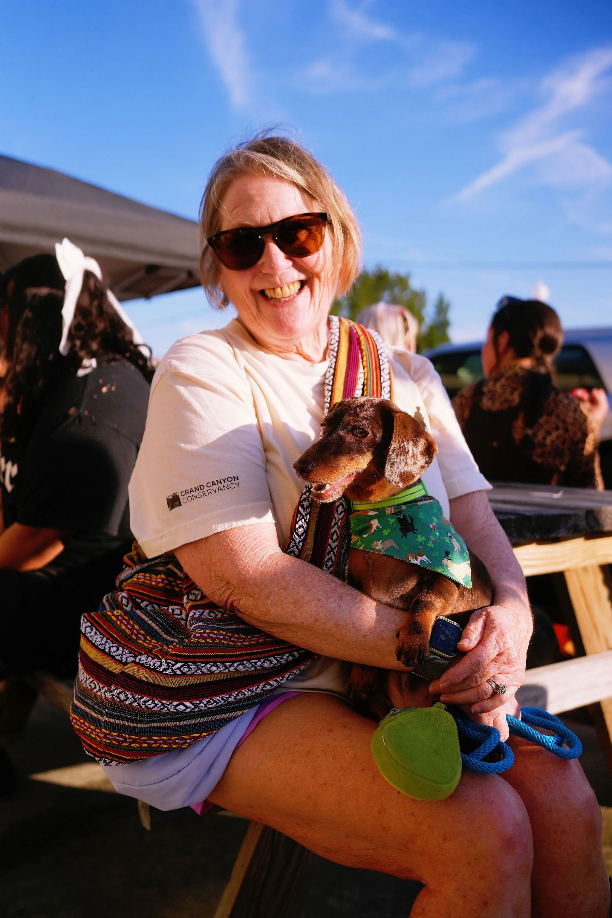 A smiling woman wearing sunglasses and a white t-shirt holding a small brown dachshund dog with a green bandana in her arms, outdoors under a blue sky with a few clouds.