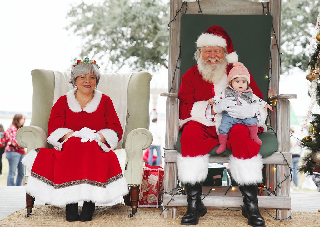 Santa Claus holding a young girl dressed in winter clothing, sitting on his lap. A woman dressed as Mrs. Claus is sitting on an armchair beside them. Christmas decorations and a Christmas tree are visible nearby.