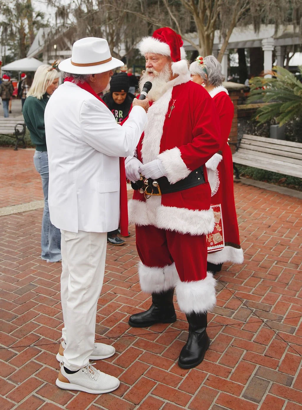 Man dressed as Santa Claus being interviewed with a microphone by a man in white suit and hat in an outdoor plaza during Christmas season.