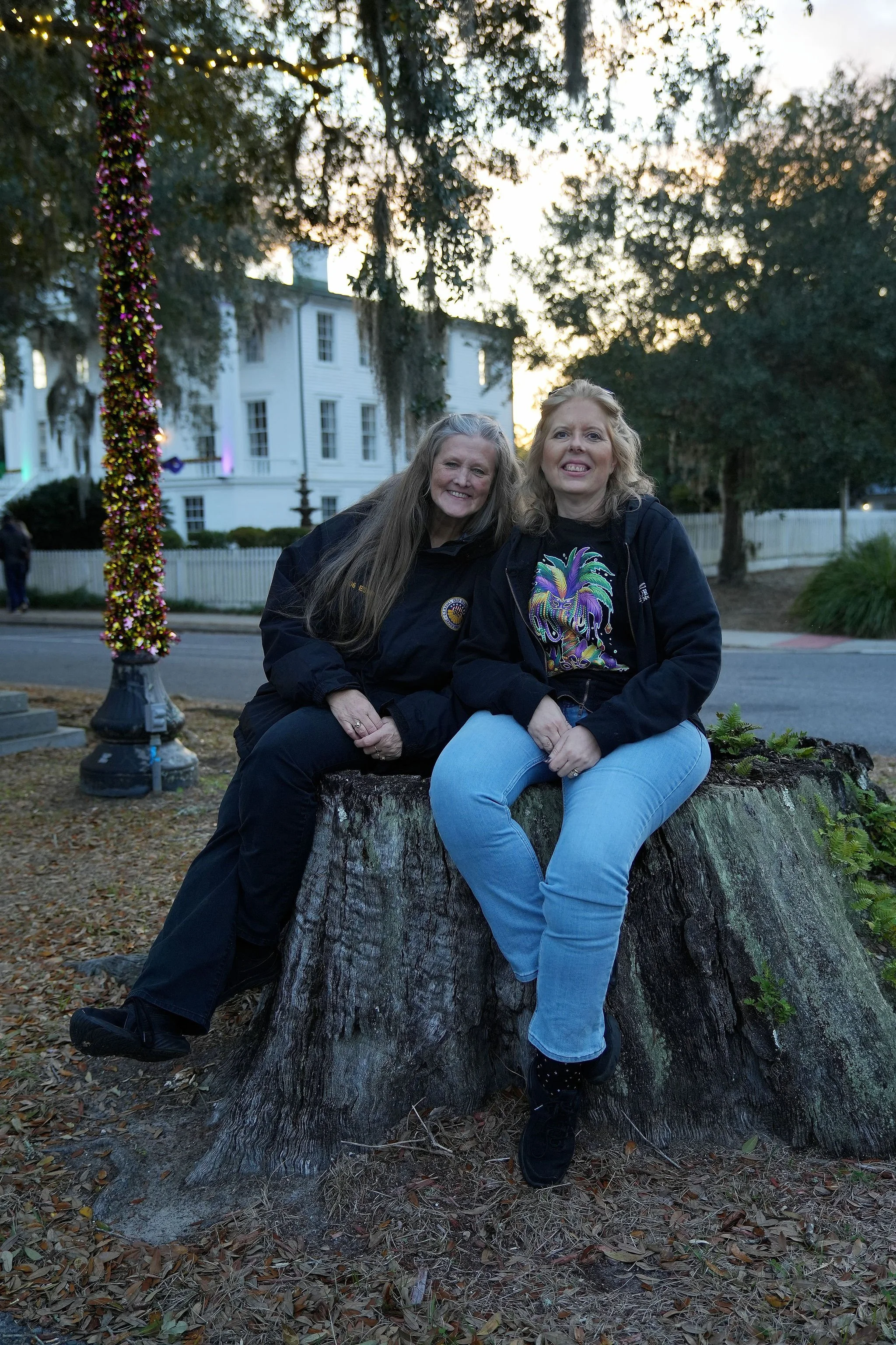 Two women sitting on a large tree stump outdoors, smiling at the camera during dusk, with trees and a white building in the background.