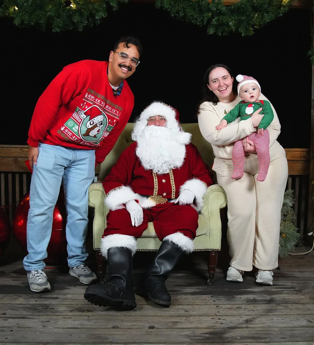 A family posing with Santa Claus, a man with a mustache wearing a red Christmas sweater, a woman holding a baby, all smiling on a wooden deck decorated with Christmas greenery.