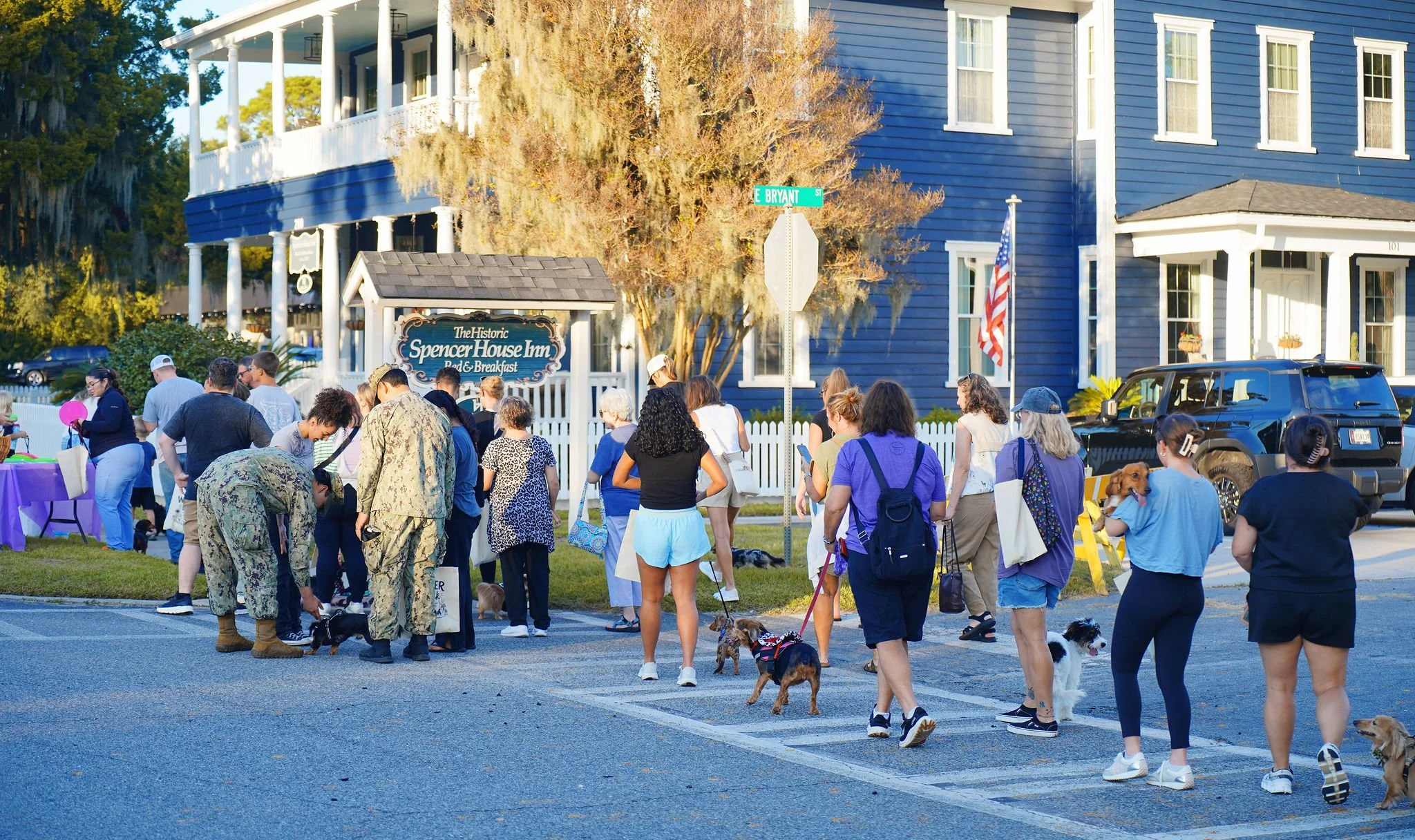 A group of people and dogs gathered outside in front of a blue building with a sign that reads 'The Historic Spencer House Inn Bed & Breakfast.' The scene appears to be a community event or gathering.