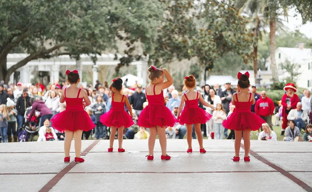 Five young girls in red ballet costumes and matching headbands performing on an outdoor stage, with a large audience seated on grass and standing in the background.