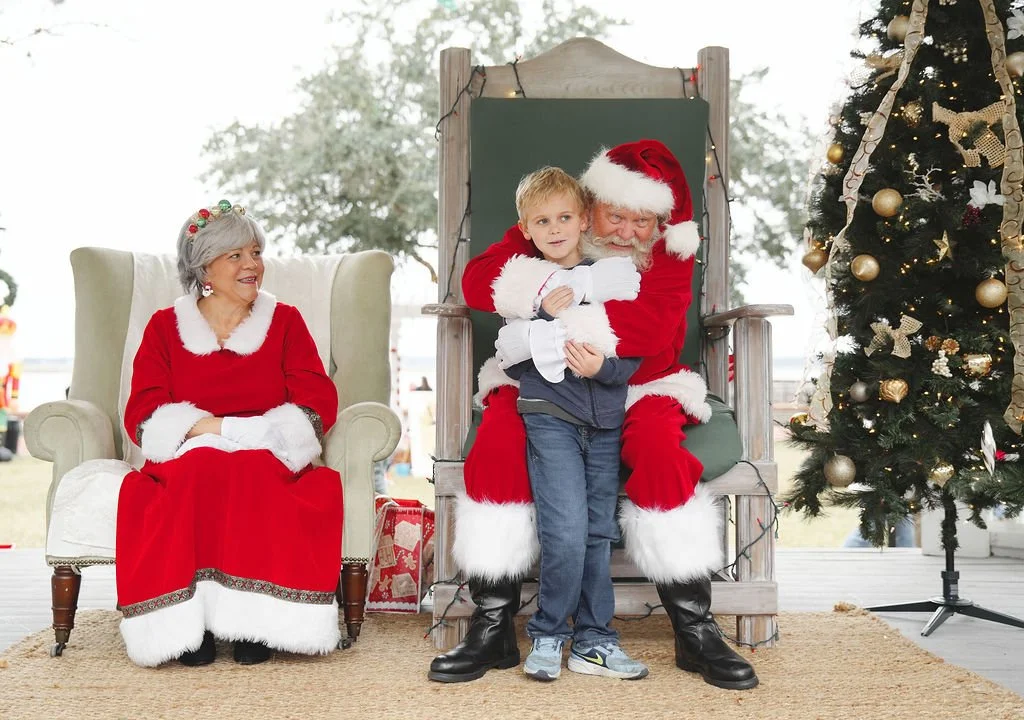 A young boy sitting on Santa Claus's lap with an older woman dressed as Mrs. Claus sitting nearby during a Christmas event.