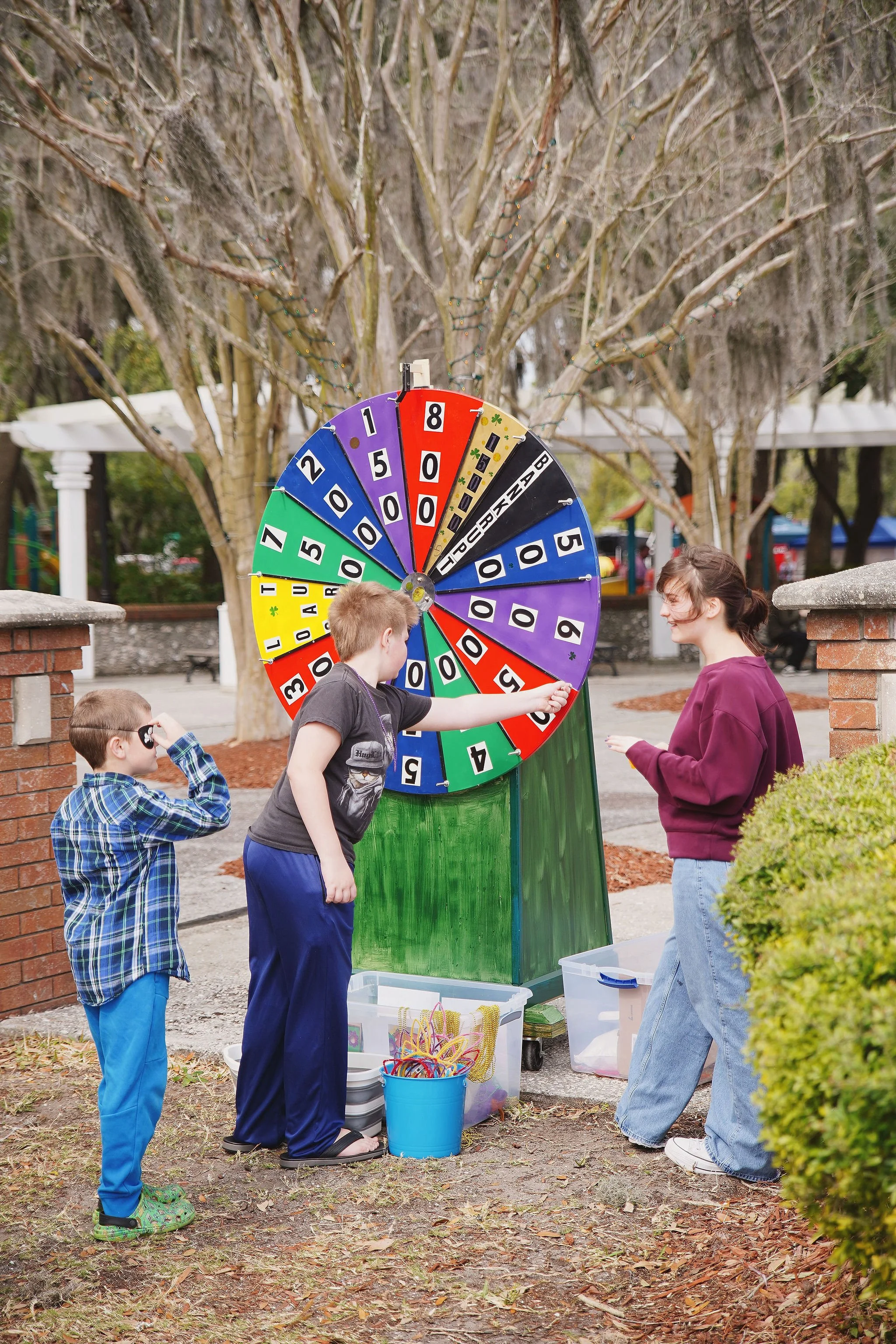 Three children stand around a colorful spinning wheel game outdoors at a park, with trees and park structures in the background.