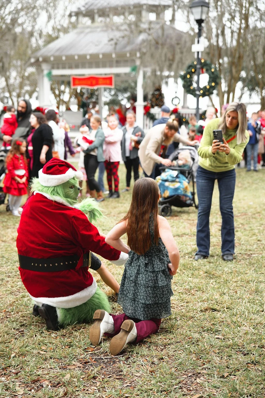 A person dressed as the Grinch, wearing a Santa hat and red velvet suit with white trim, kneeling on the grass and holding a girl's hand. The girl has long brown hair, is wearing a dark dress with white shoes, and is kneeling on the grass. In the bac