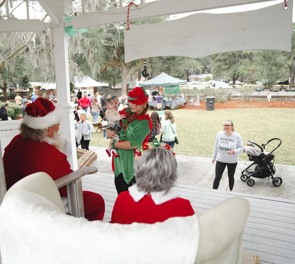 People at a Christmas event; Santa Claus, a woman dressed as an elf holding a baby, and another woman in a red and white dress sitting on a porch, with a crowd and tents in the background.