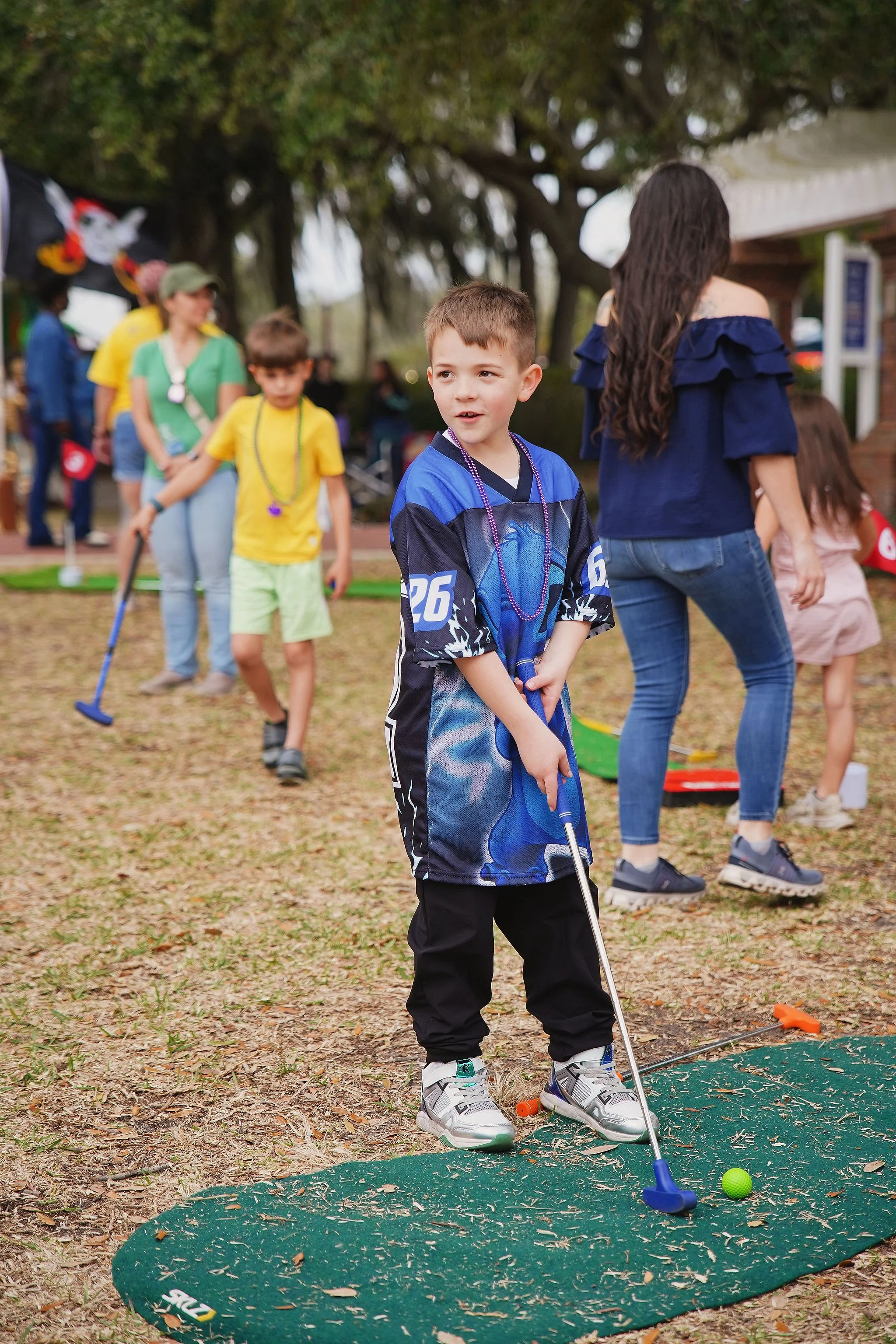 A young boy in a blue sports jersey is holding a golf club, standing on a green mat, preparing to hit a green golf ball. Other children and adults are in the background at an outdoor event, with trees and tents visible.