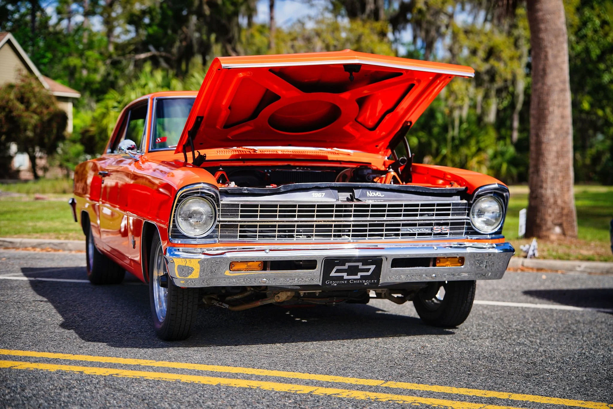 A red vintage Chevrolet Nova SS car with its hood open, parked on a street with trees and houses in the background.