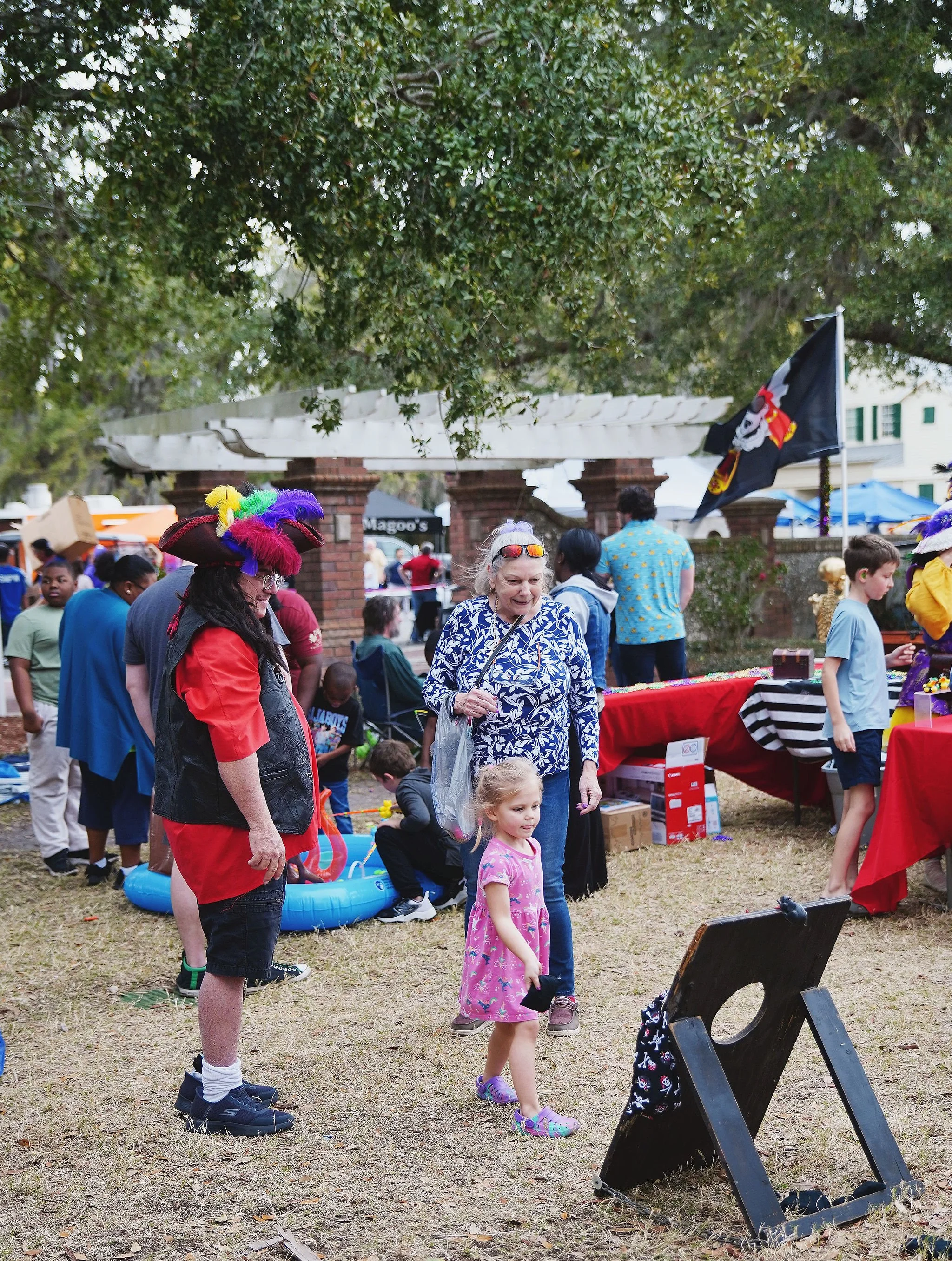People at a festival or event, with a pirate flag and various vendor tables, children playing, and some wearing colorful costumes and hats, under large trees.