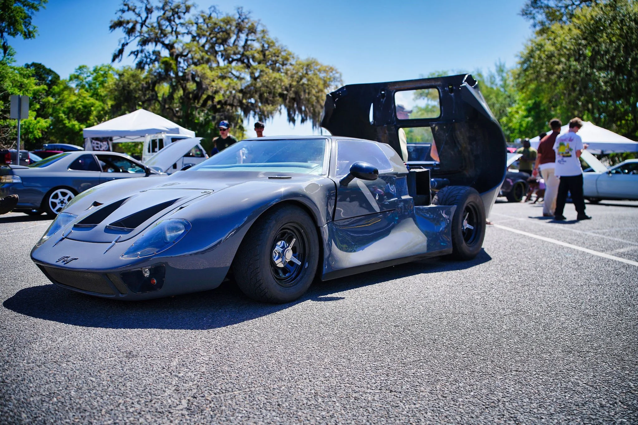 A sleek, black sports car with an open rear section at a car show, surrounded by other vehicles and people under blue skies and green trees.