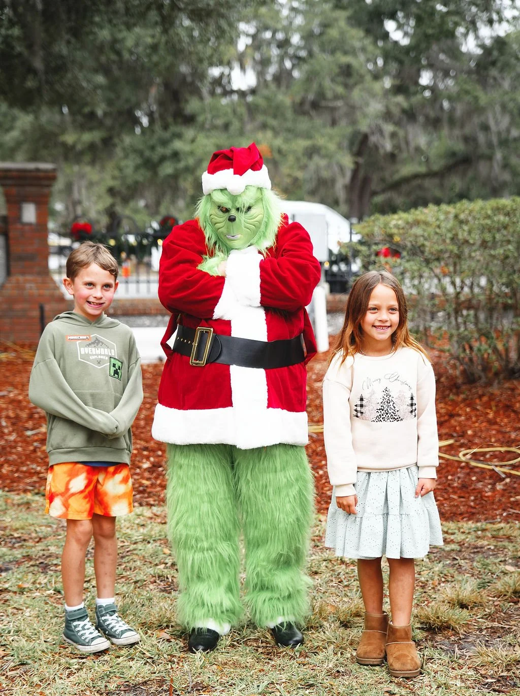 A person dressed as the Grinch in a Santa Claus outfit with a Santa hat, standing between two children outdoors during Christmas. The boy is wearing a green hoodie with a graphic, orange shorts, and sneakers. The girl is wearing a white sweater with 