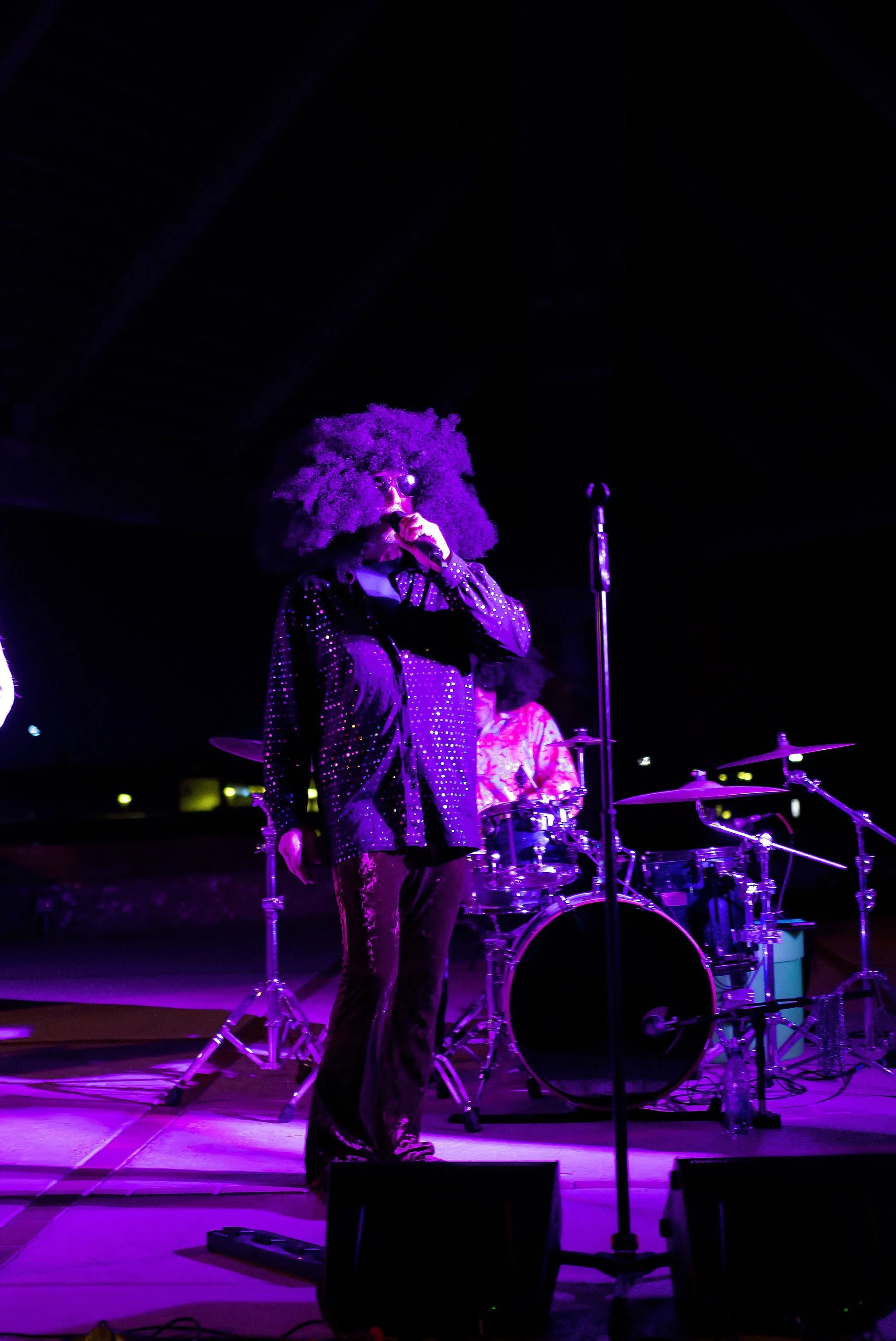 A singer with large curly hair and dark sunglasses performing on stage with purple lighting, accompanied by a drummer in the background.
