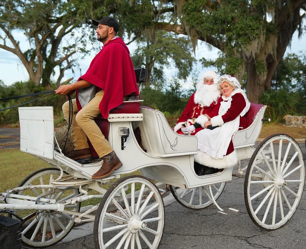 A man dressed as Santa Claus and a woman dressed as Mrs. Claus sitting in a vintage white horse-drawn carriage, being driven by a man in a red cape and cowboy hat in a park with large trees.