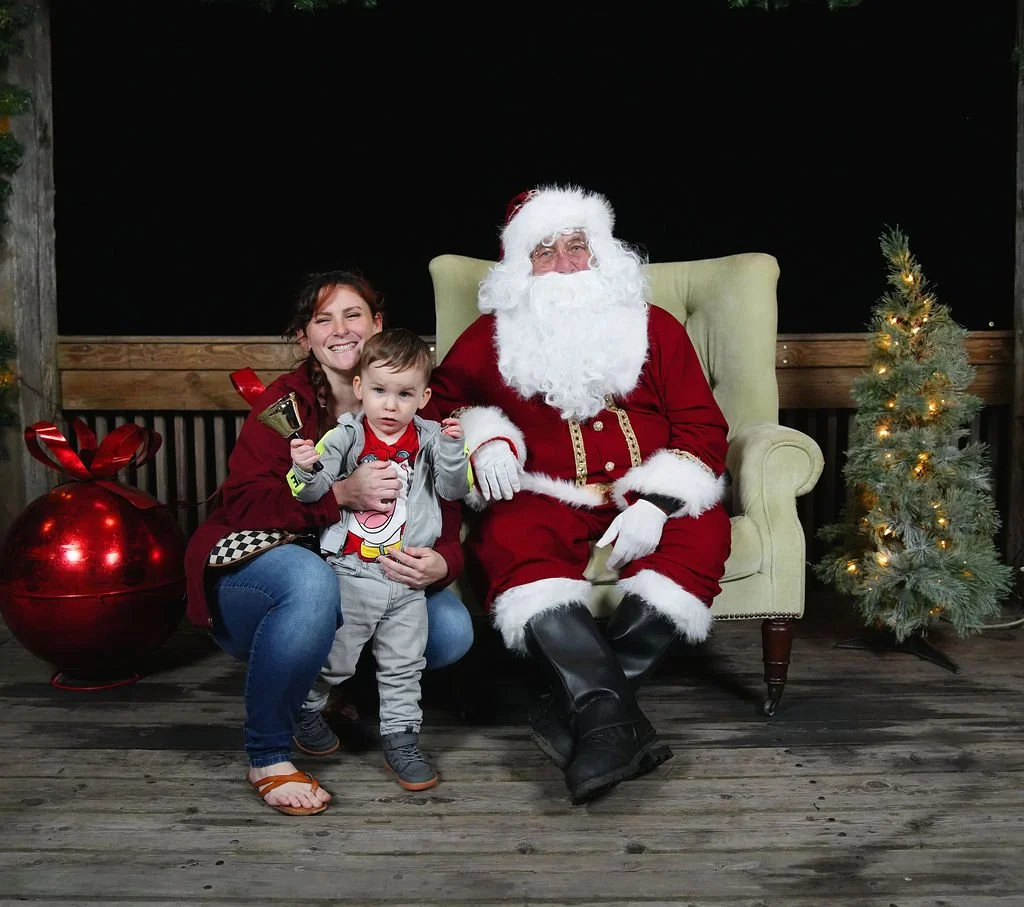 A woman, a young boy, and Santa Claus pose together during Christmas. The woman is smiling, holding the boy, who is holding a small bell, while sitting on a wooden floor. Santa is seated in a green armchair, wearing his traditional red suit and white