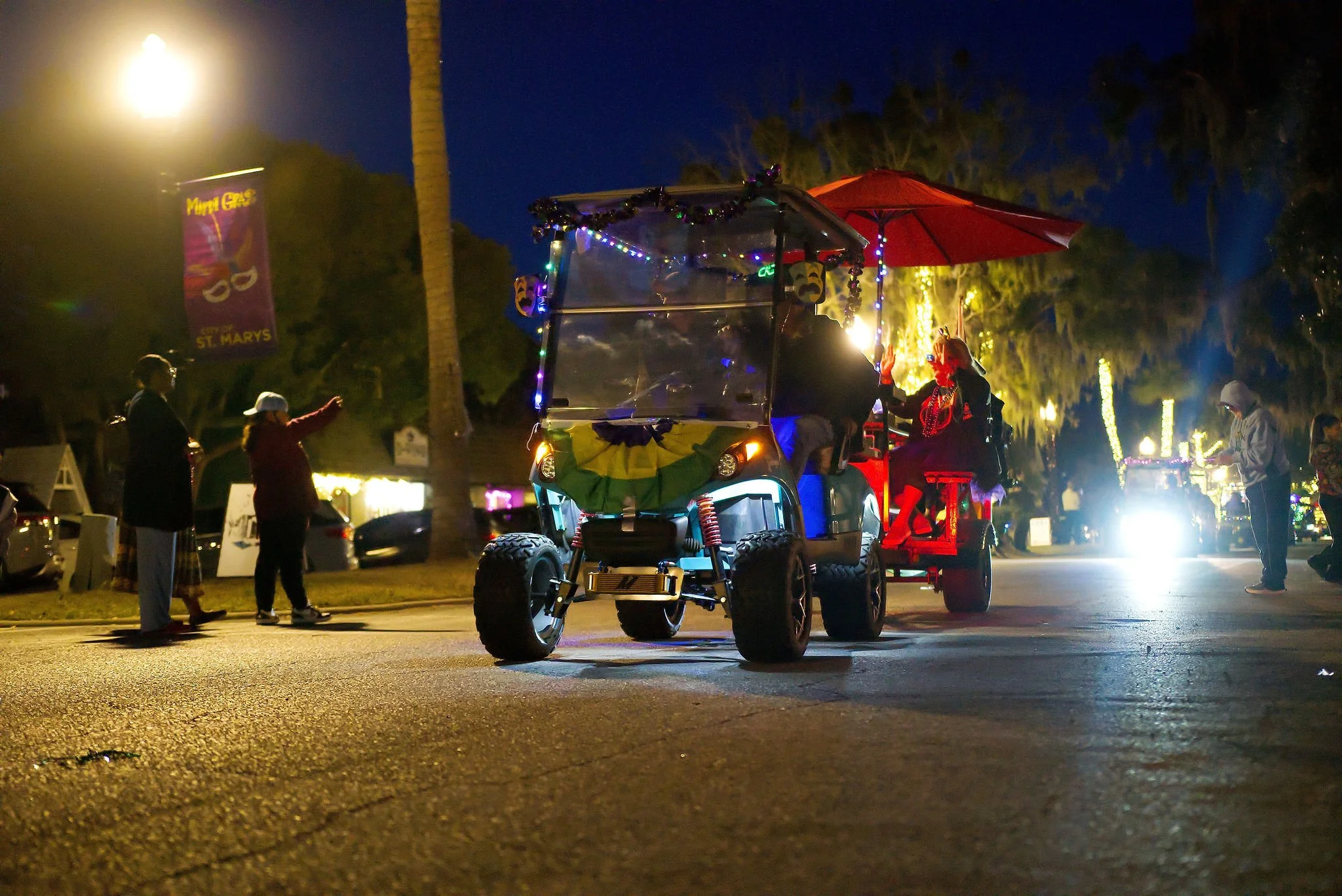 A nighttime parade featuring decorated golf carts, with people dressed in costumes and accessories, illuminated by streetlights and festive lights in the background.