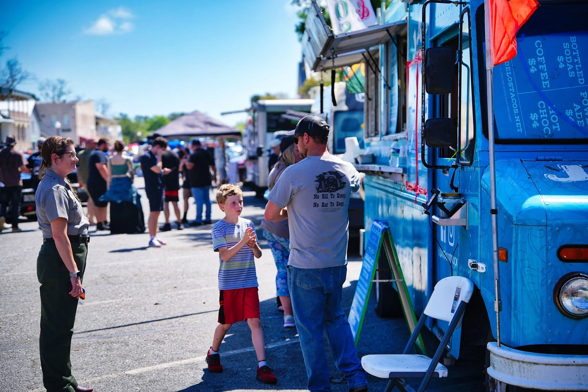 People at a farmer's market or food truck fair on a sunny day, including a woman in uniform, a young boy, and a man at a food truck.
