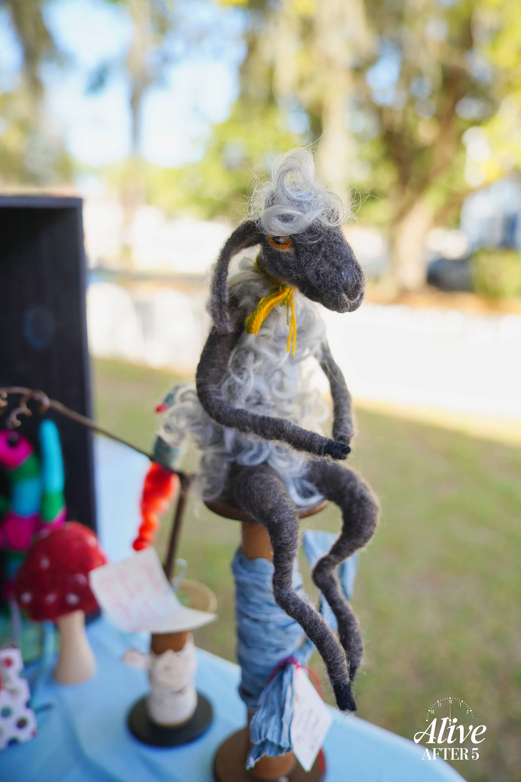 Felted stuffed horse doll with curly hair, seated on a stand, outdoors with trees in the background.
