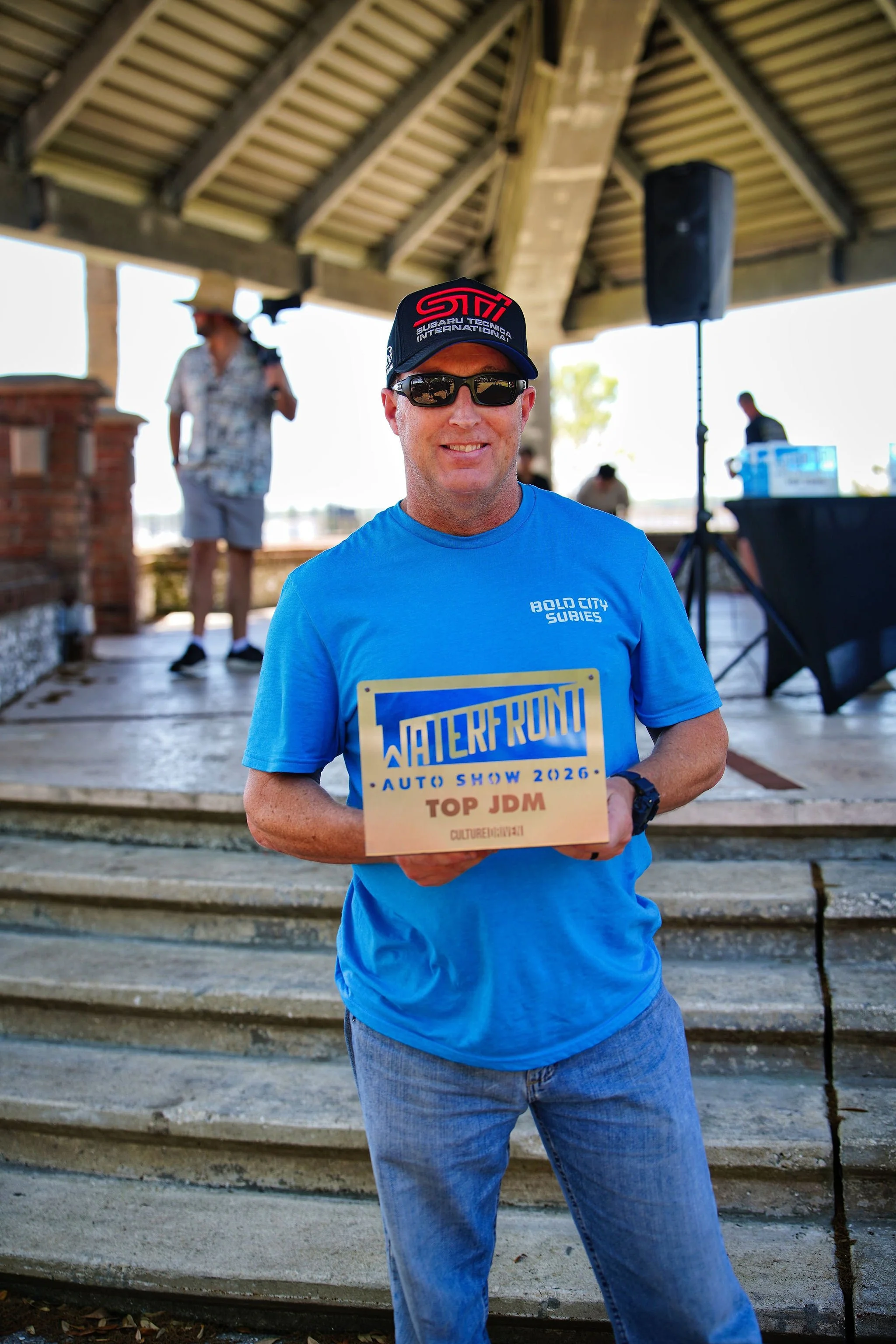 A man in a blue t-shirt and sunglasses holding a sign, standing on steps at an outdoor event. The sign reads 'WATERFROUNT AUTO SHOW 2026, TOP JDM.' Under a pavilion, with other people and a speaker in the background.