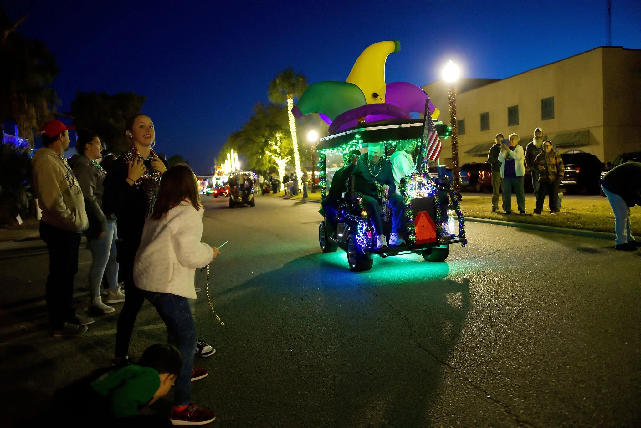 Nighttime parade scene with a decorated golf cart with colorful balloons and holiday lights, and people watching along the street.