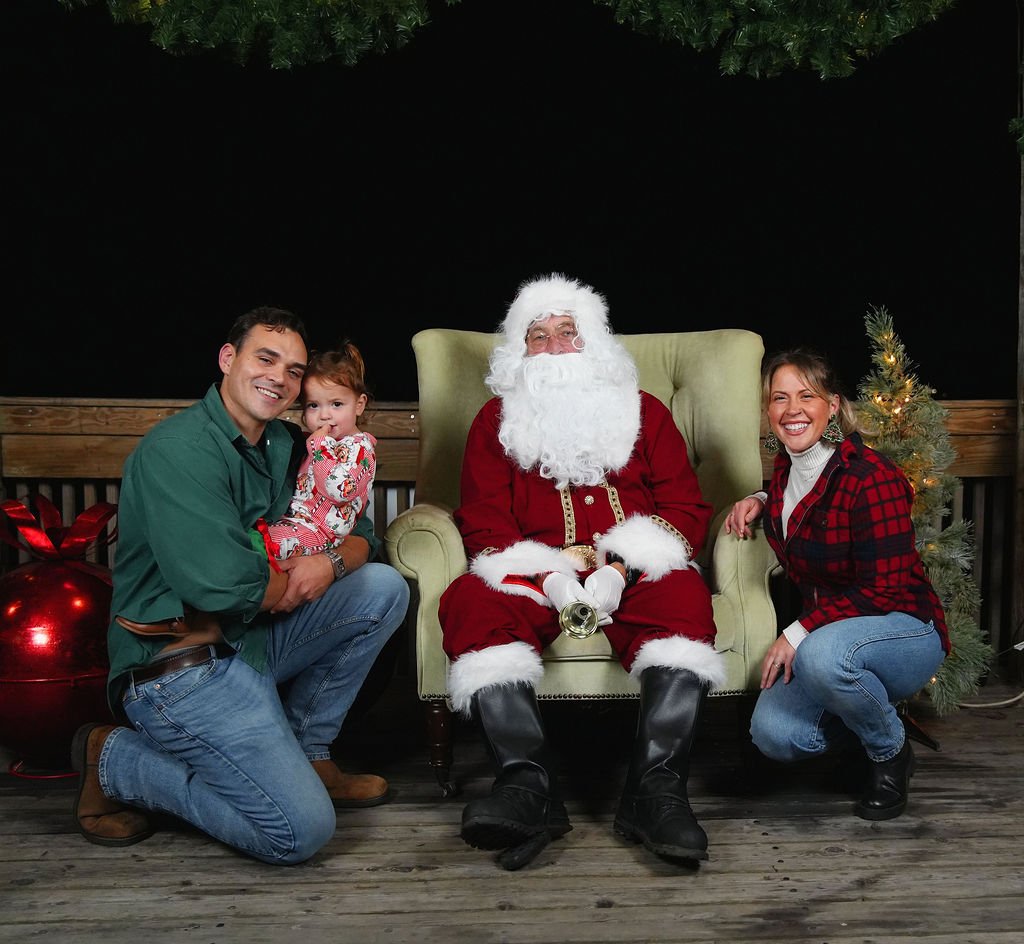 Family posing with Santa Claus on a wooden porch decorated for Christmas, including a small Christmas tree and ornaments.