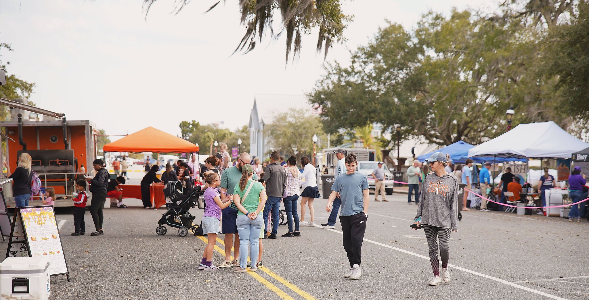 People walking and browsing at an outdoor street fair with vendor tents and food stands under trees on a cloudy day.