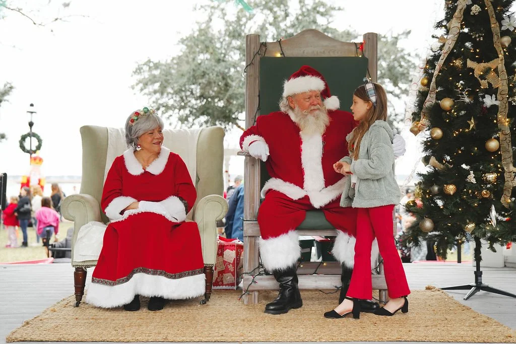 A young girl talking to Santa Claus who is sitting on a throne with Mrs. Claus sitting nearby, at a Christmas event outdoors.