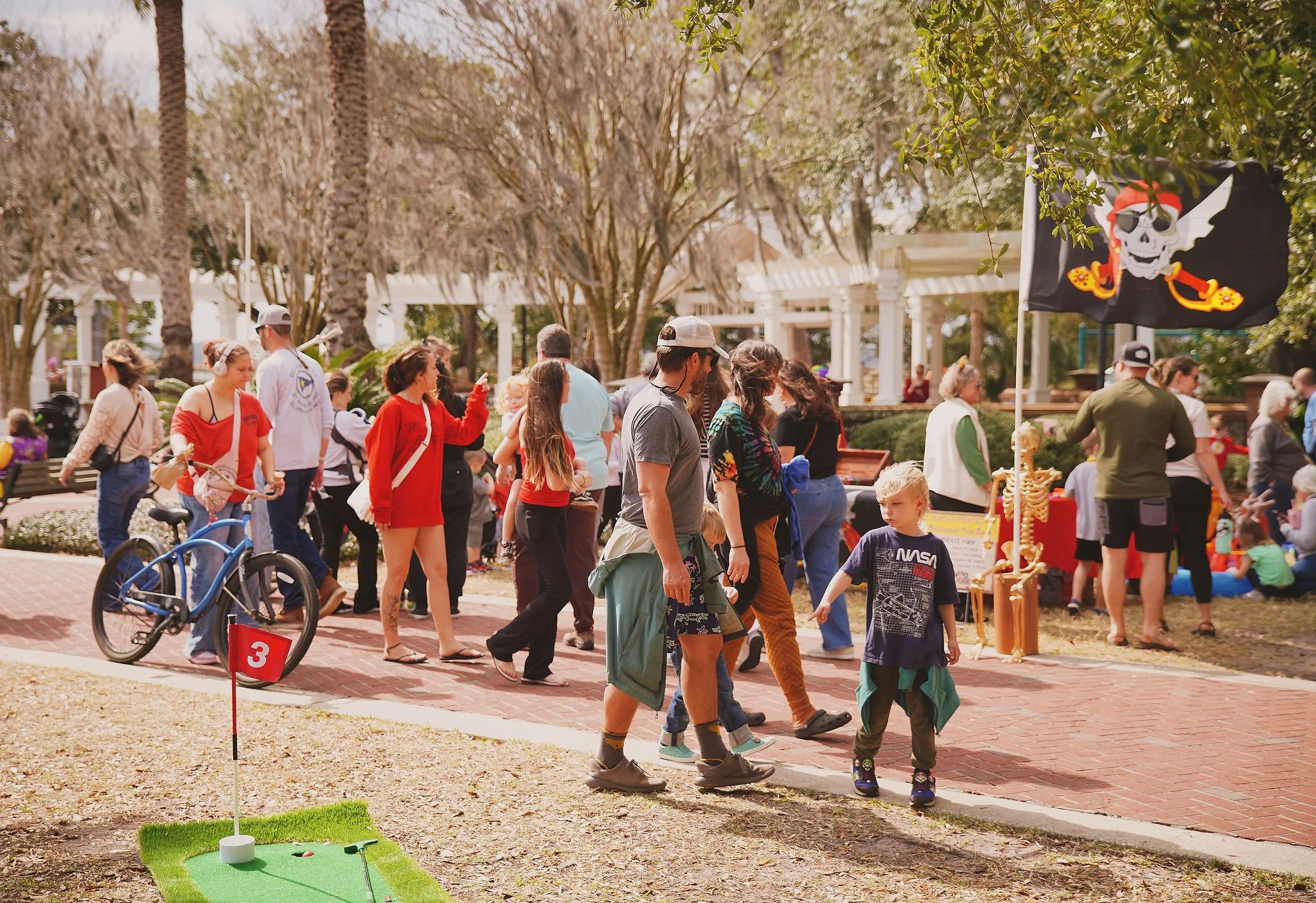 People outdoors at a pirate-themed event with a pirate flag displaying a skull with an eye patch and a pirate hat, mini golf setup on the grass, and trees in the background.
