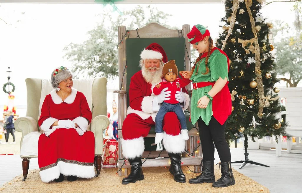 A young girl dressed as an elf interacts with a baby sitting on Santa's lap, while Santa and a woman dressed as Santa's wife look on during a Christmas event with a decorated Christmas tree nearby.