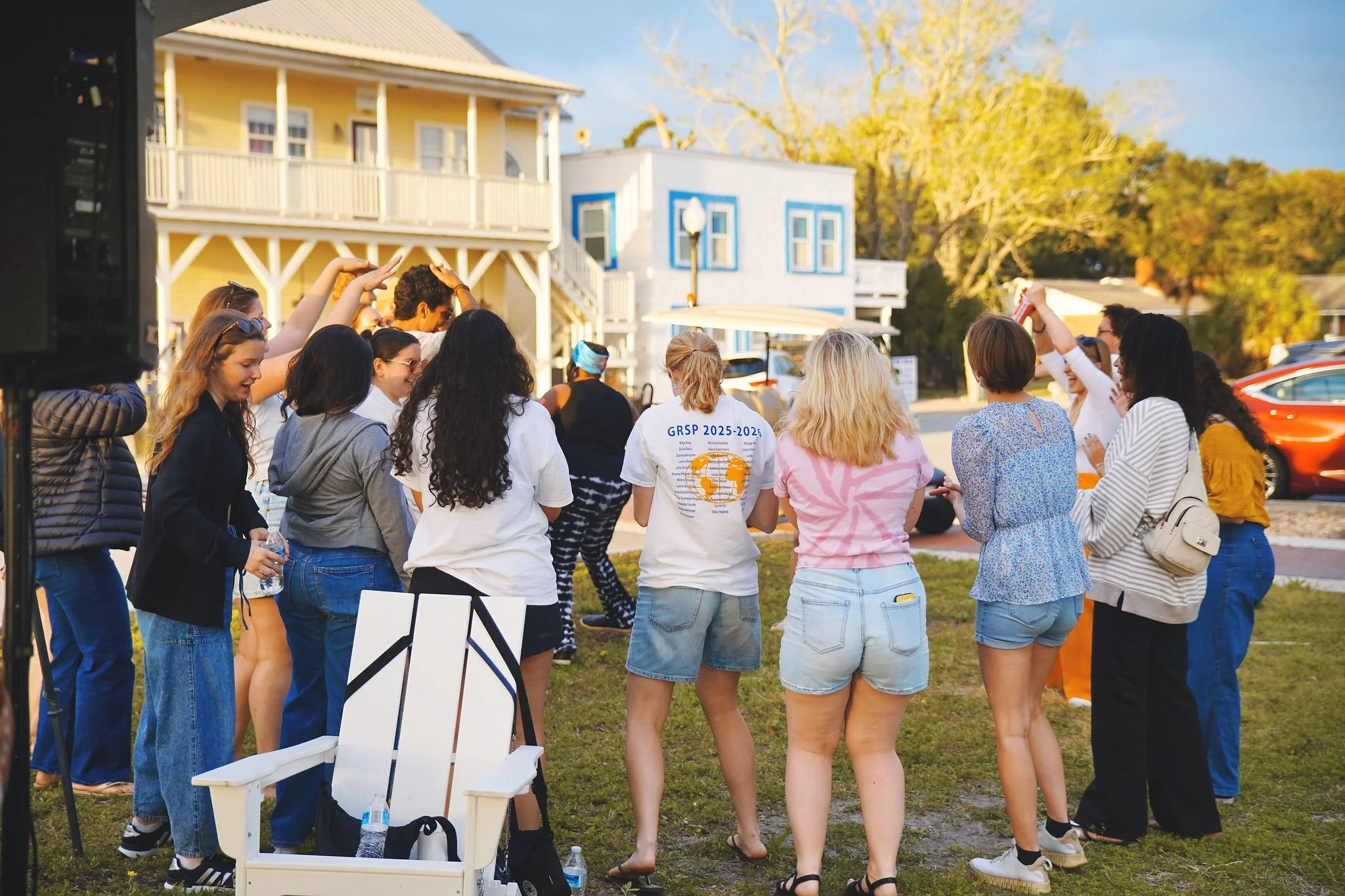 Group of people gathered outdoors on a lawn, dancing and socializing during a sunny day, with colorful houses and trees in the background.