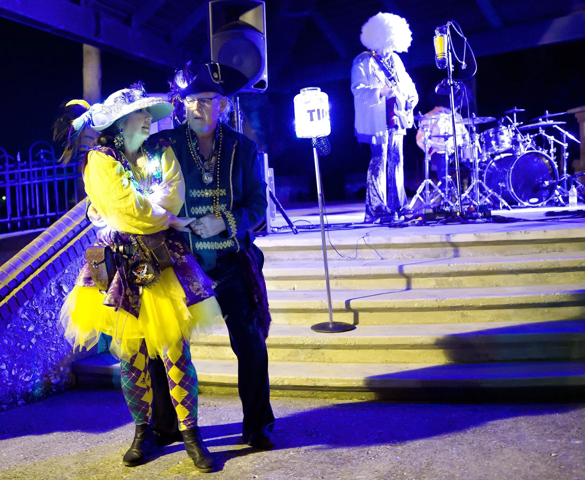 Two performers dressed as pirates dancing in front of a stage with a band and drum set, illuminated by colorful stage lights at night.