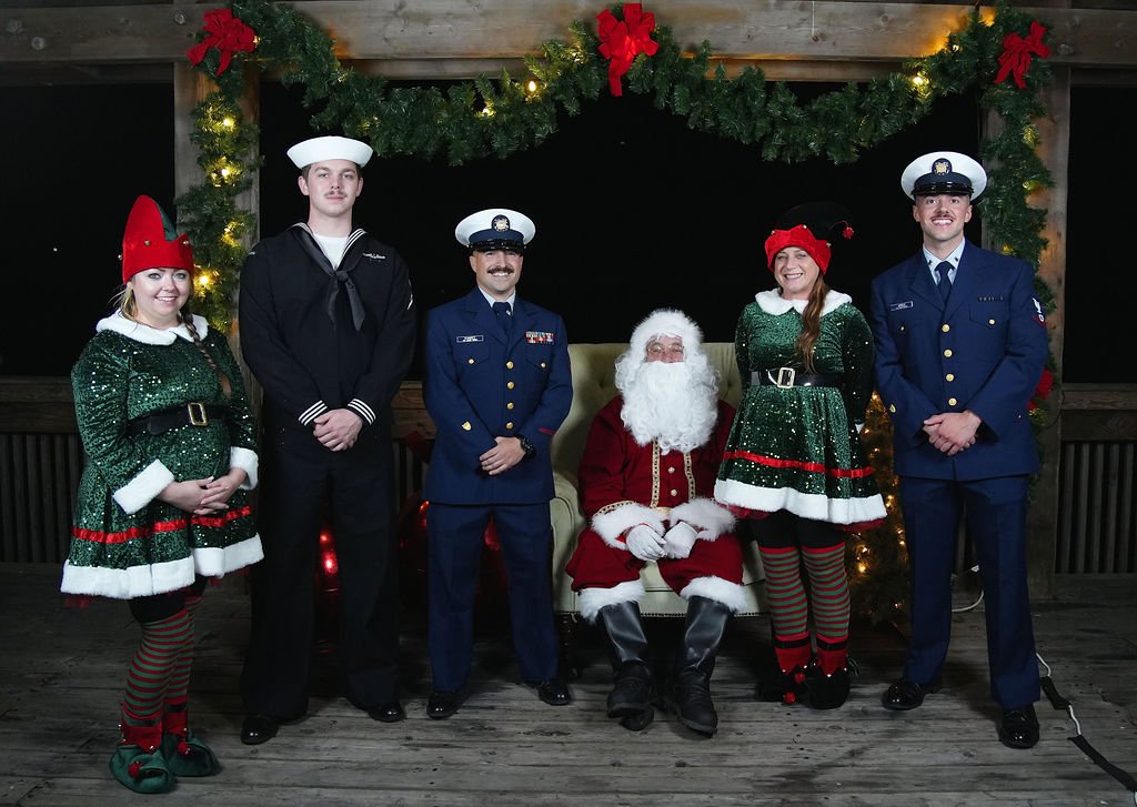 Group of people dressed in Christmas costumes, with Santa Claus seated in the center, flanked by three people dressed as military or navy personnel, and two women dressed as elves, standing on a decorated porch at night.