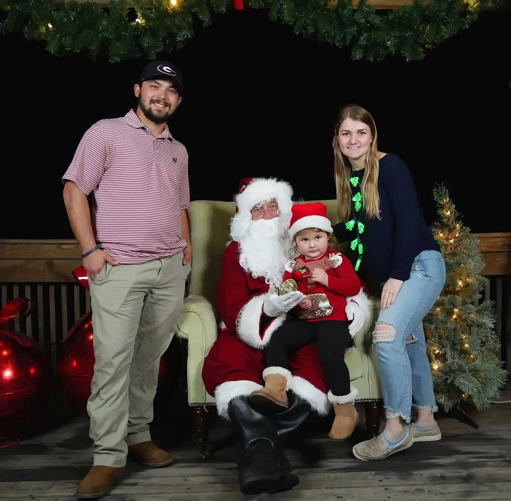 Four people posing with Santa Claus on a Christmas-themed set. Santa is sitting in a green chair, holding a young girl wearing a red Christmas dress and hat, who is holding a small wrapped gift. Two adults, a man in a striped shirt and beige pants, a