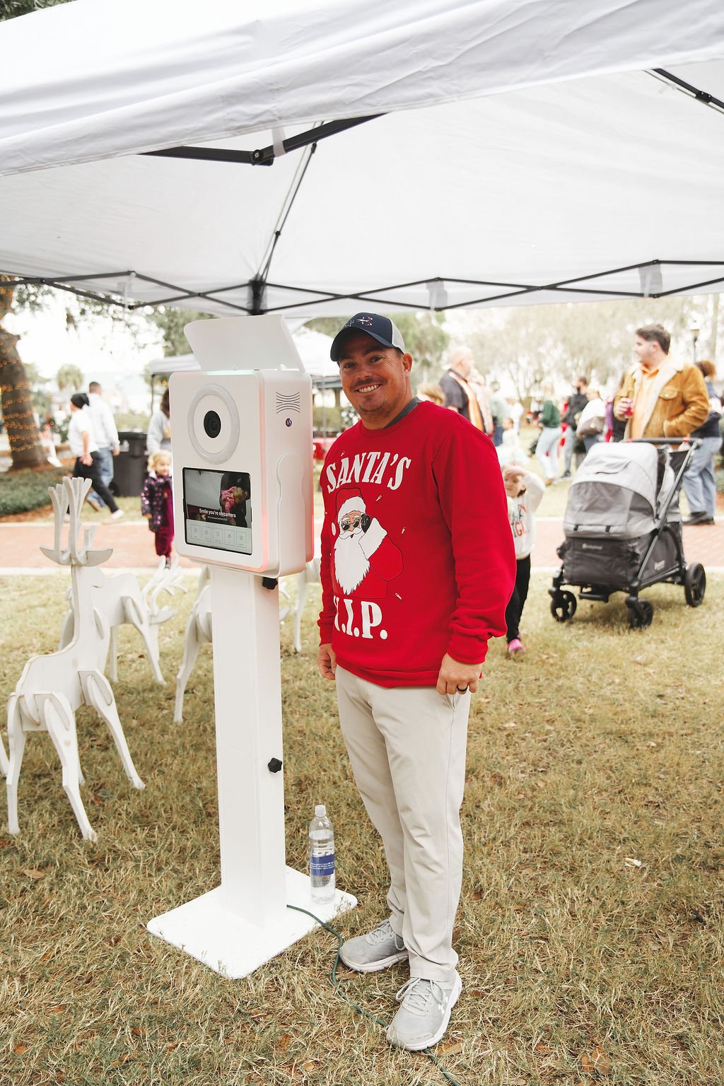 Man smiling at camera under a white tent, wearing a red Christmas sweater with Santa and 'R.I.P.' on it, standing next to a white photo booth with a screen and camera, on a grassy area with Christmas decorations and people in the background.