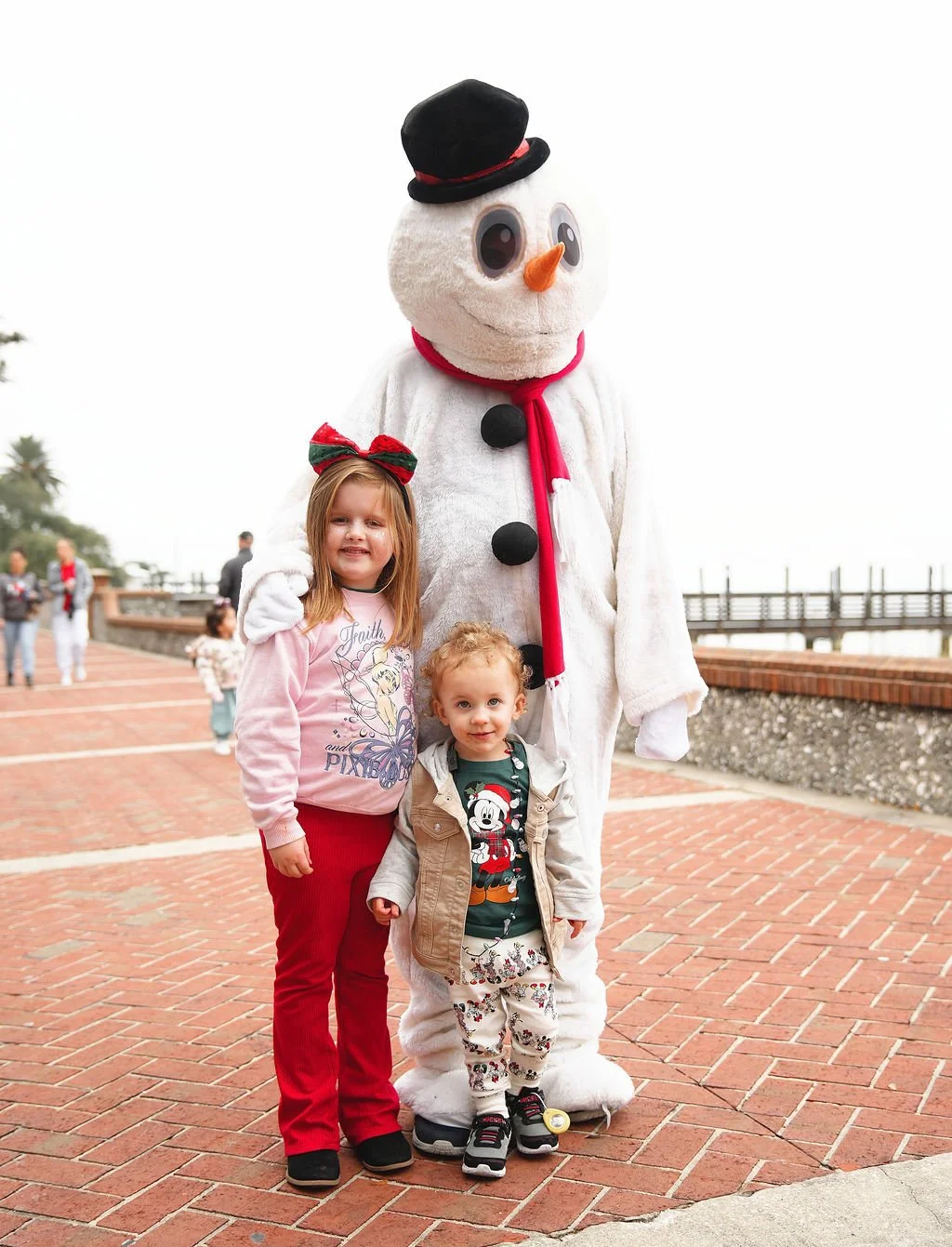 Two young children standing next to a person in a snowman costume with large eyes, an orange carrot nose, and black buttons, wearing a black top hat and red scarf. The scene appears to be outdoors on a brick-paved walkway during daytime.